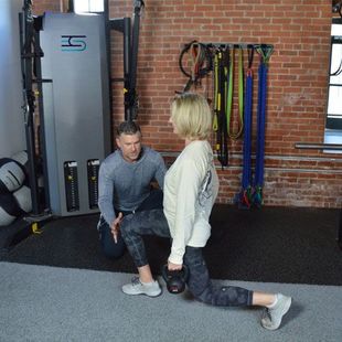 A person performing a lunge exercise, guided by a trainer in a gym.
