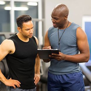 Two men are standing next to each other in a gym looking at a tablet.