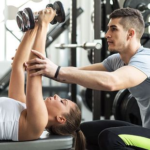 A man is helping a woman lift a dumbbell in a gym.