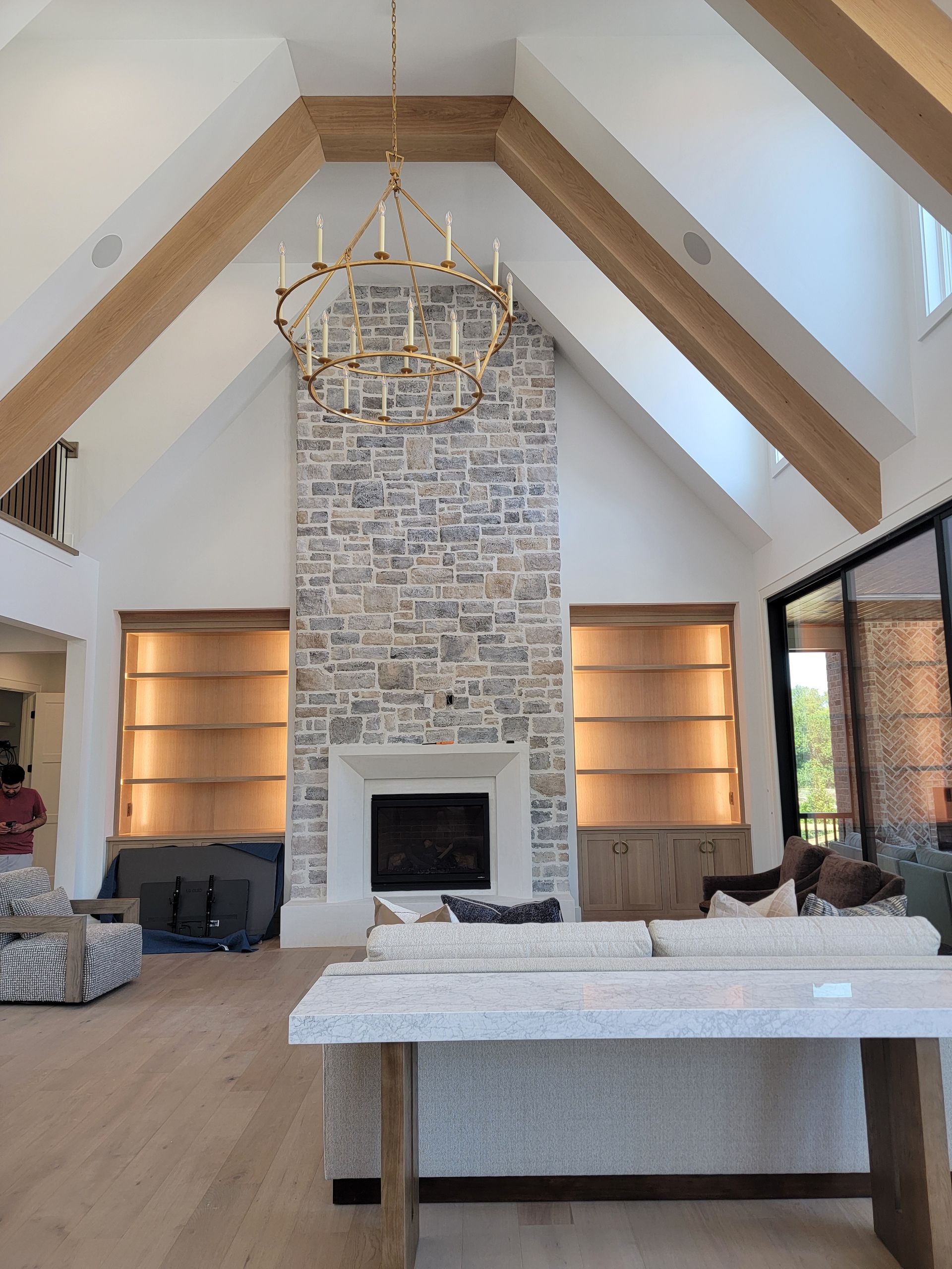 Living room with stone fireplace, wood beams, chandelier, built-in shelves, and large glass doors.