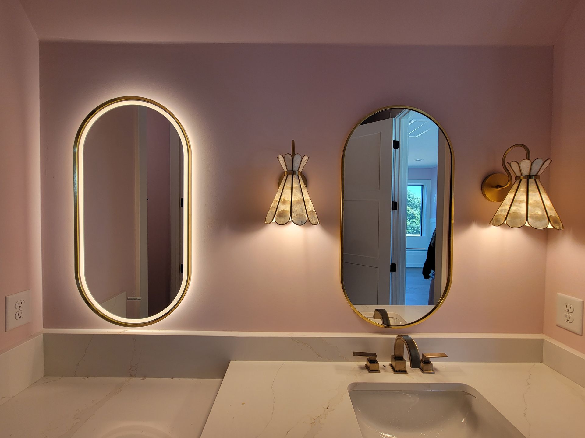 Pink bathroom with gold-framed oval mirrors, sconces, and a white sink.