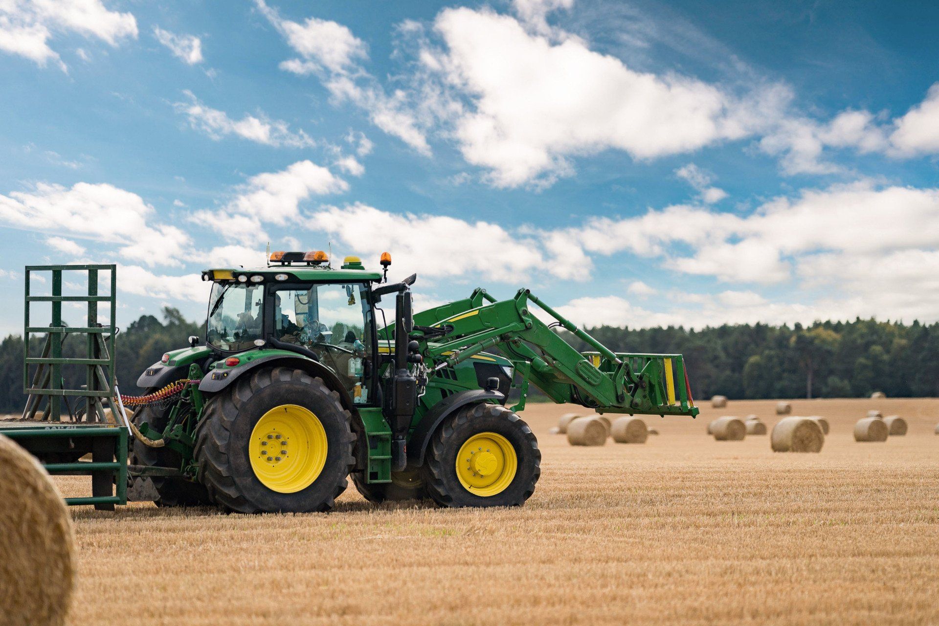 Green tractor with loader in a field of hay bales under a blue sky.