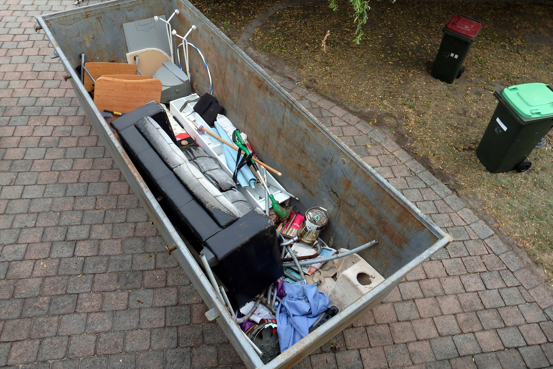 A dumpster filled with junk is sitting on a brick sidewalk.