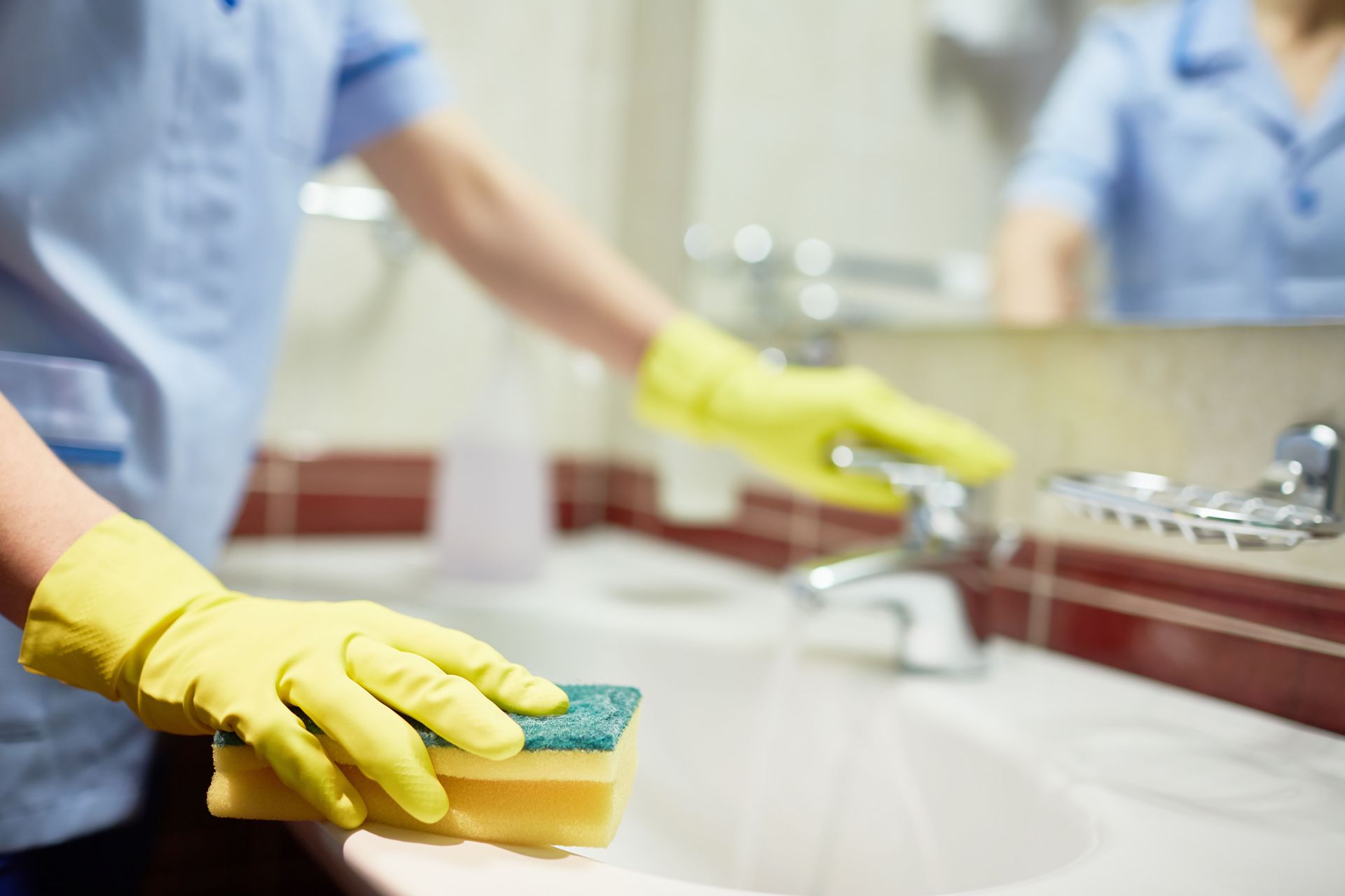 A person wearing yellow gloves is cleaning a bathroom sink with a sponge.
