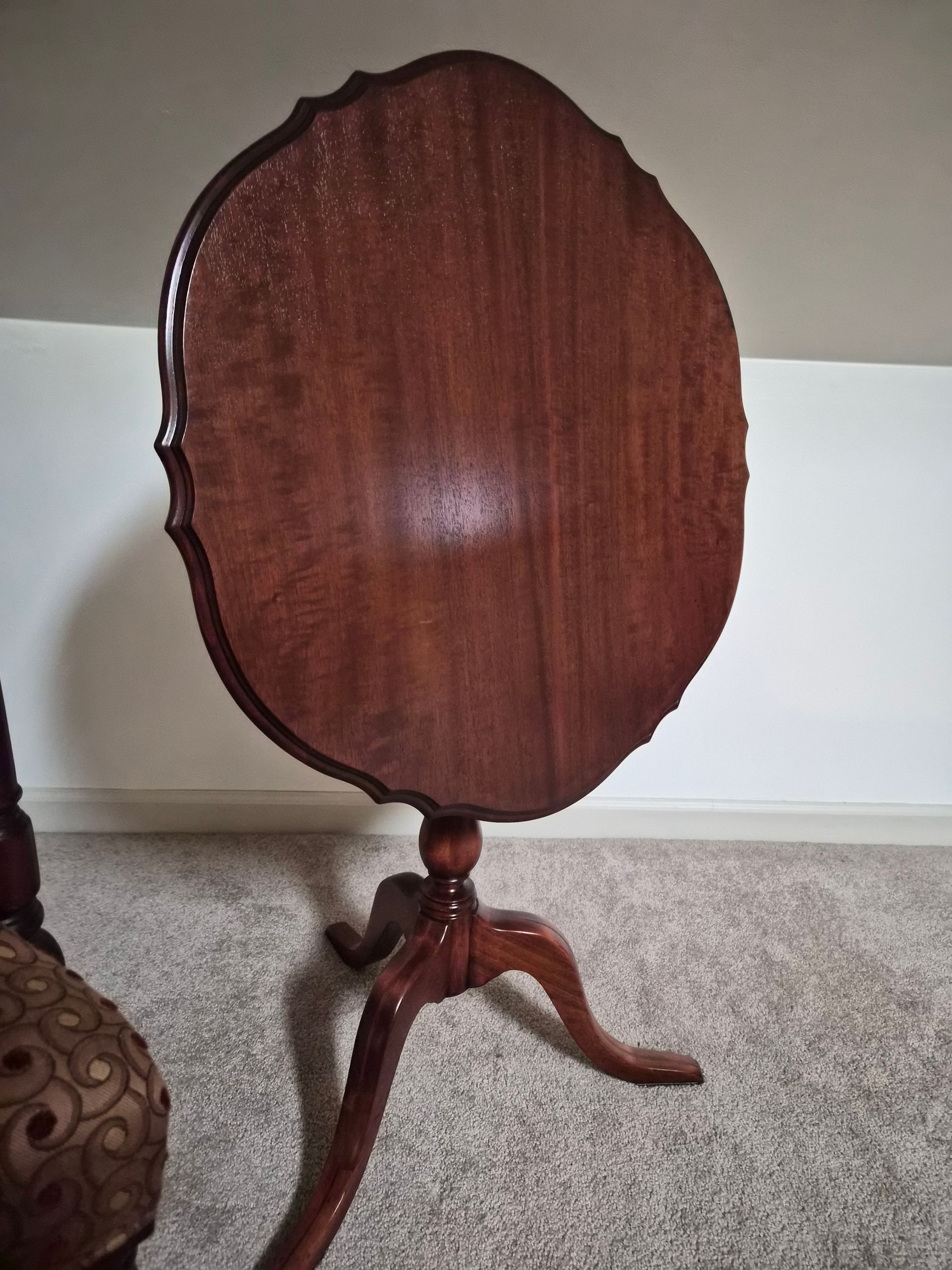Brown wooden tilt-top table with scalloped edge, three legs, on a carpeted floor.