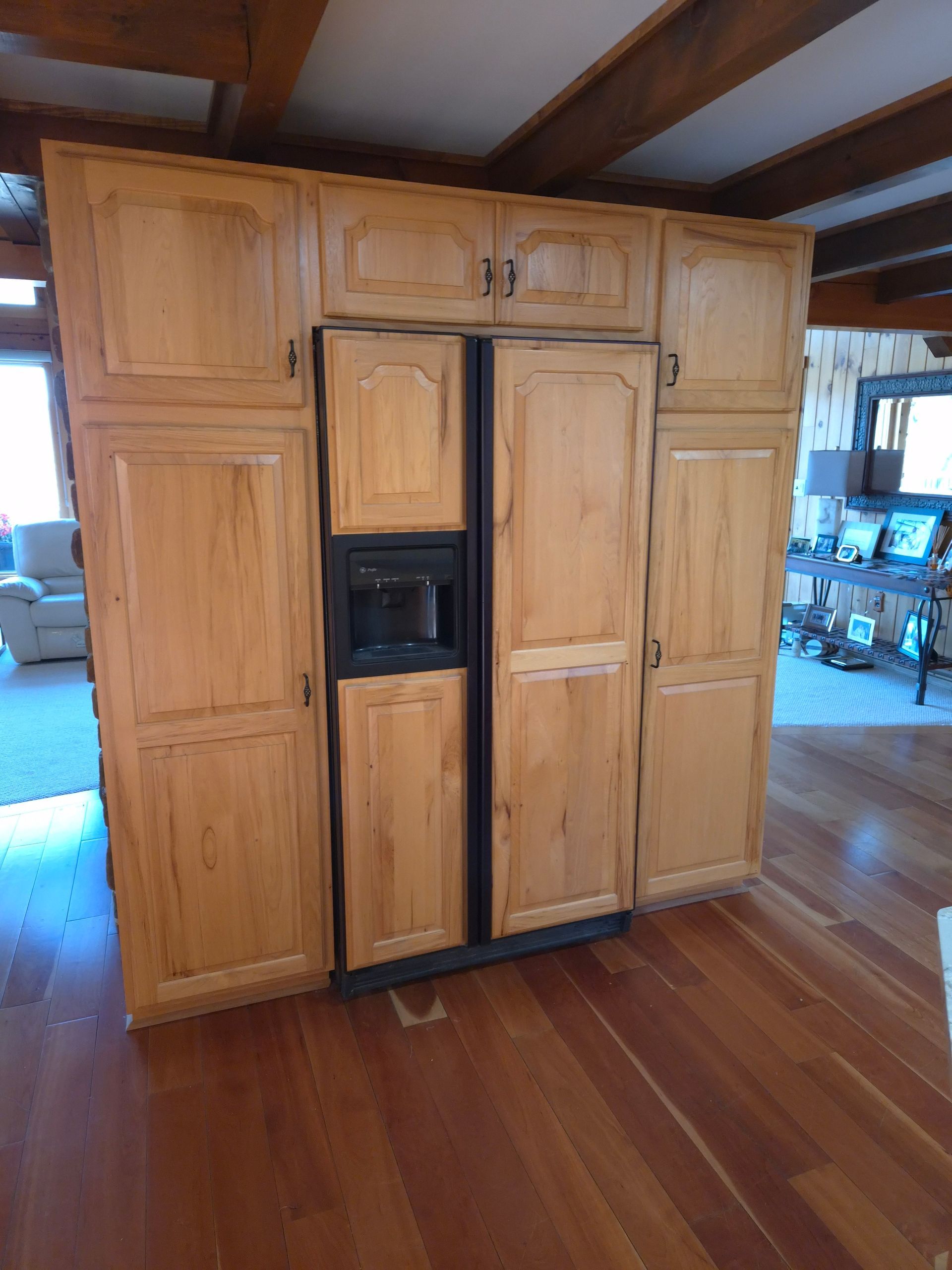 Light wood kitchen cabinets surround a black refrigerator. Wooden floor in foreground.
