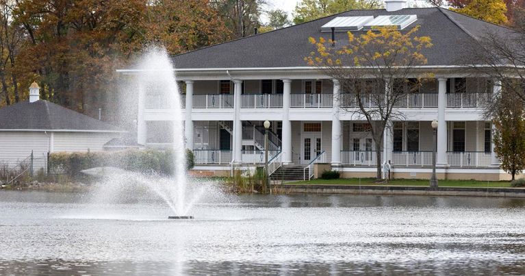 A large white house is sitting next to a lake with a fountain in front of it.