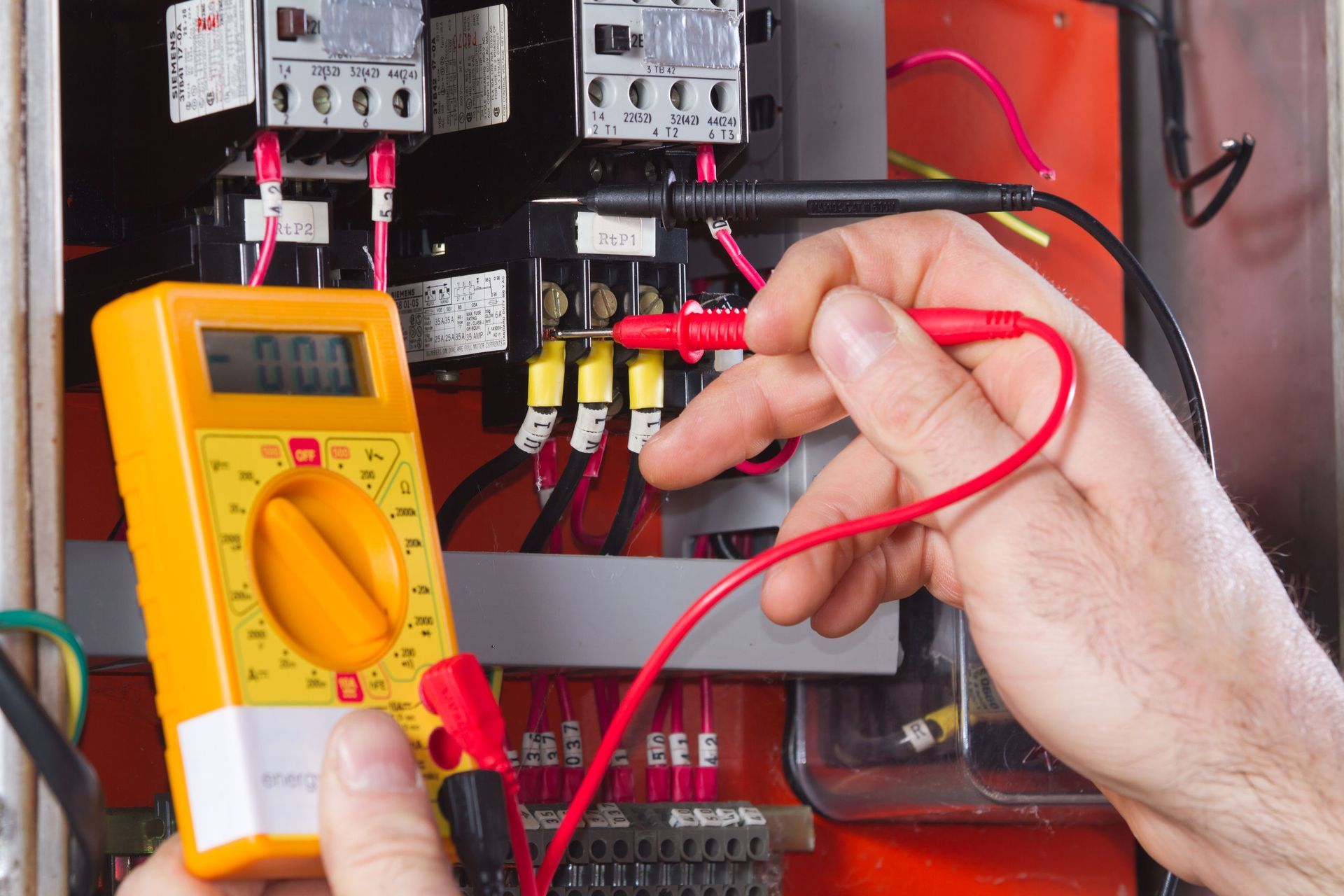 Hand testing an electrical panel with a yellow multimeter and red probes