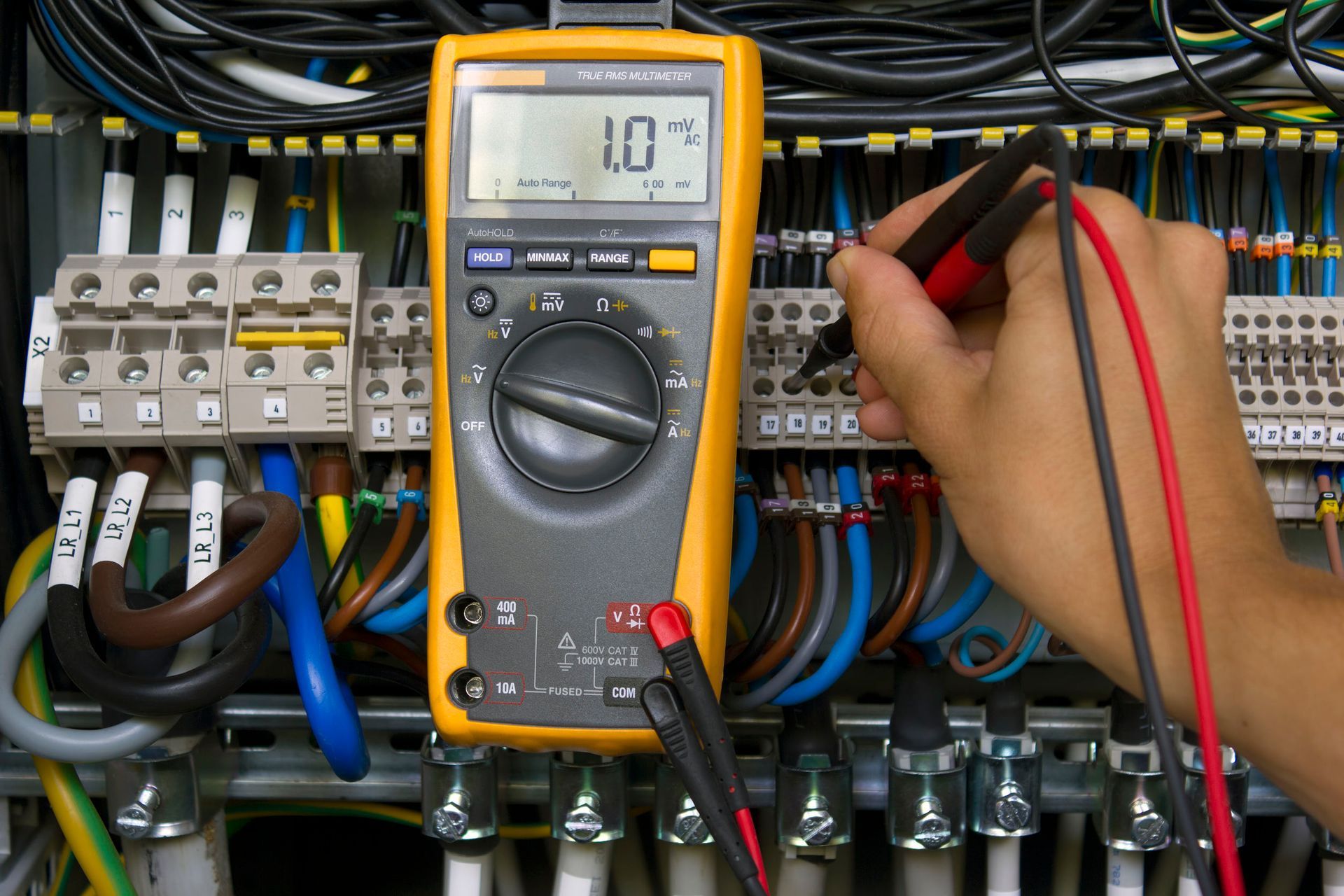 Digital multimeter testing wiring in an electrical control panel with bundled cables and circuit breakers