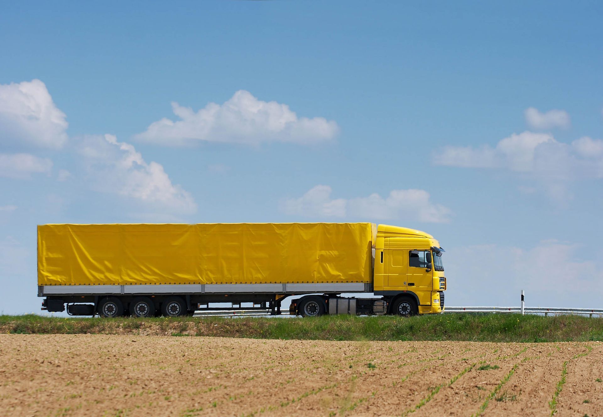 Yellow semi-truck with a covered trailer on a road, blue sky with clouds in the background.