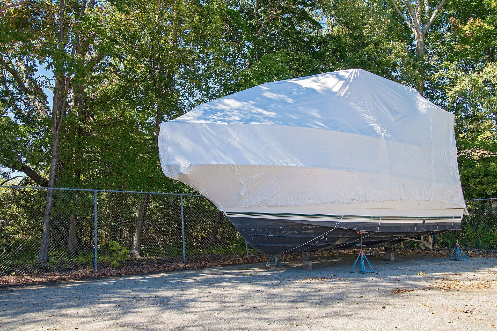 Boat covered in white tarp, parked near trees behind a fence.