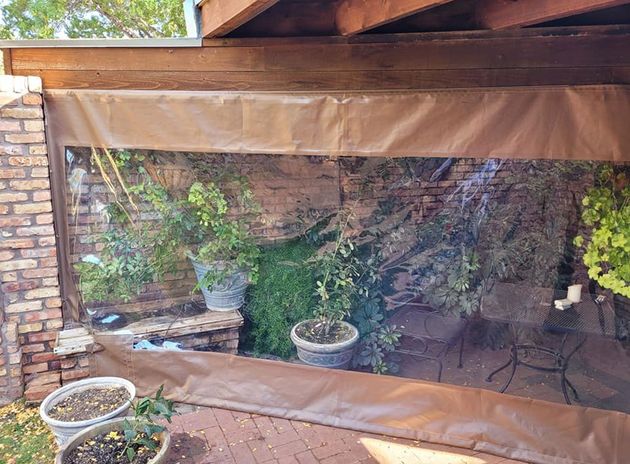 Clear vinyl outdoor shade, brown fabric trim, plants visible through, brick wall.