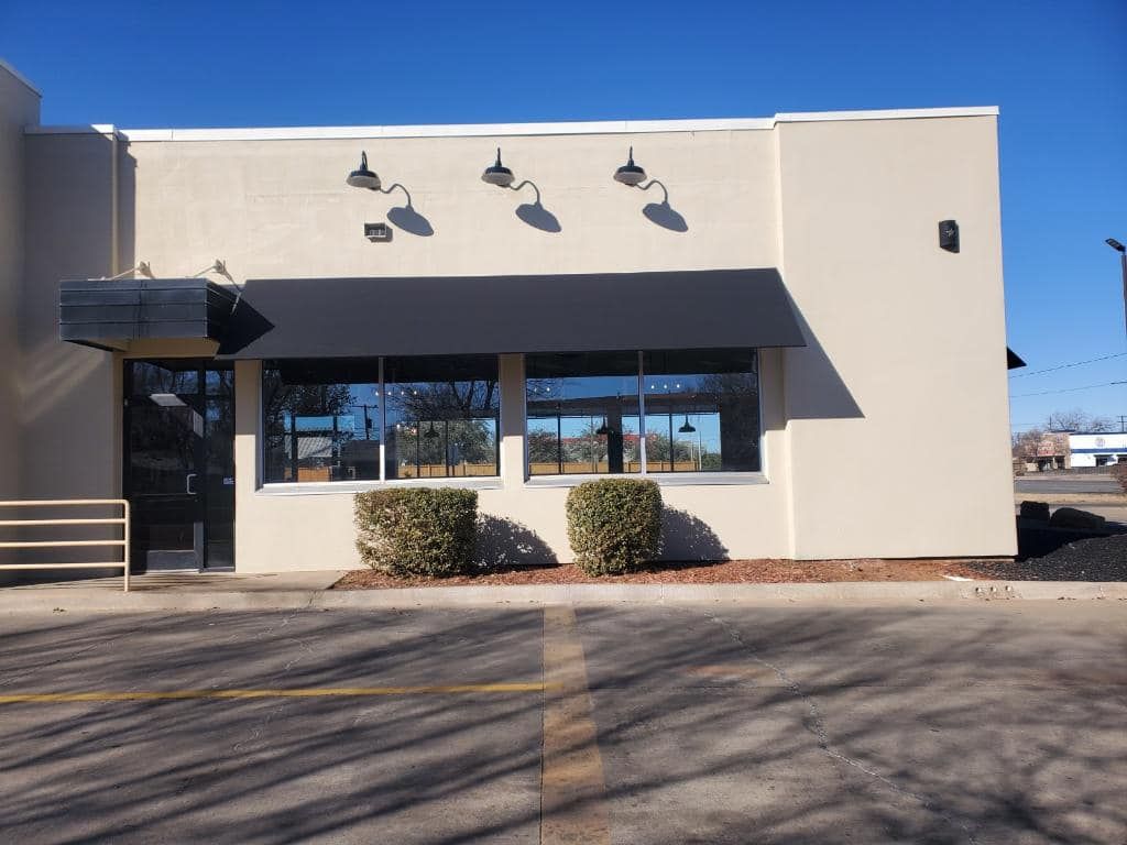 Exterior of a business with a black awning and windows. Beige walls, bushes in front.