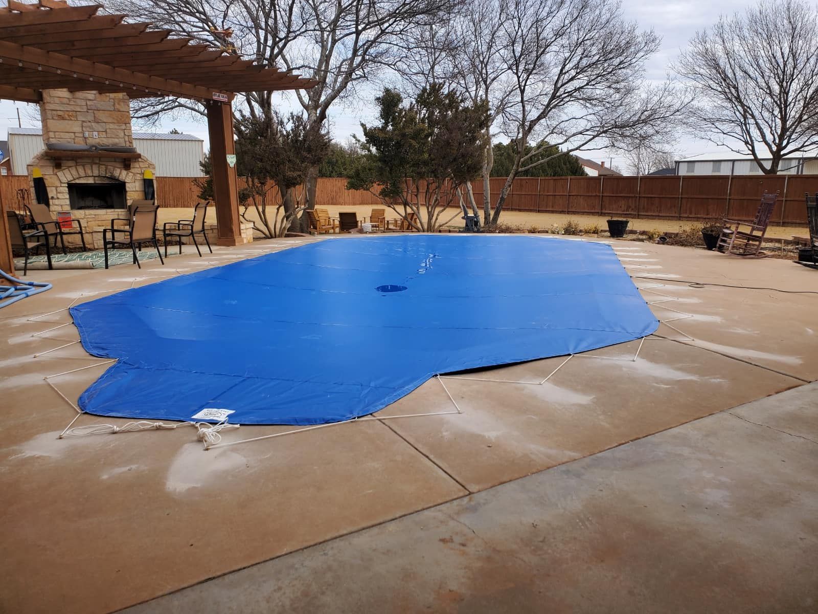 A blue pool cover on a concrete patio, with a pergola and outdoor fireplace visible in the background.