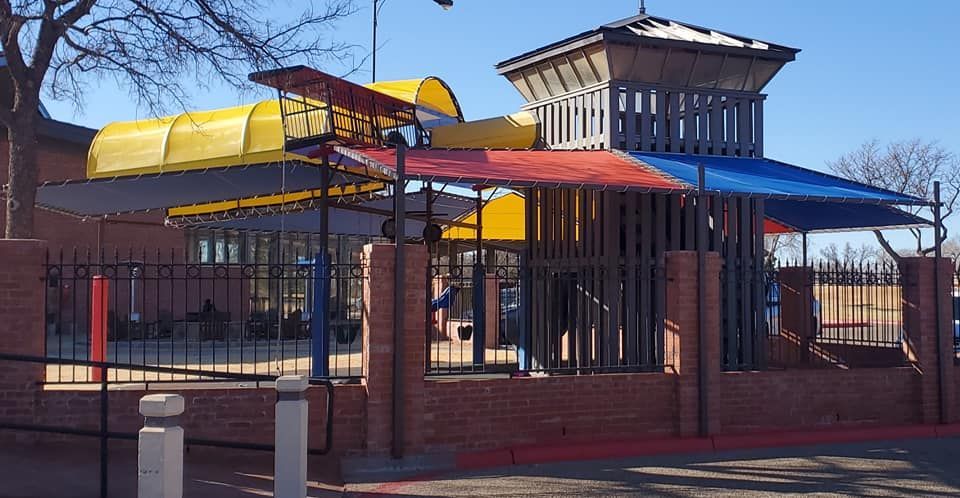 Playground with yellow, red, and blue shades, brick walls, and a black fence.