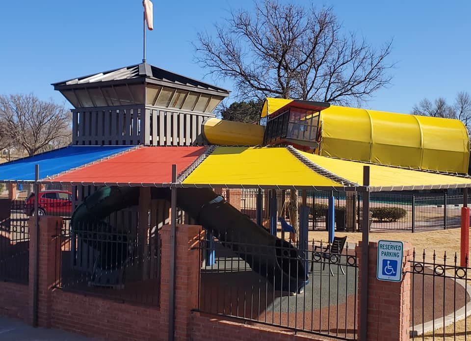 Playground with colorful sunshades, a tower, and a slide behind a brick and wrought-iron fence.