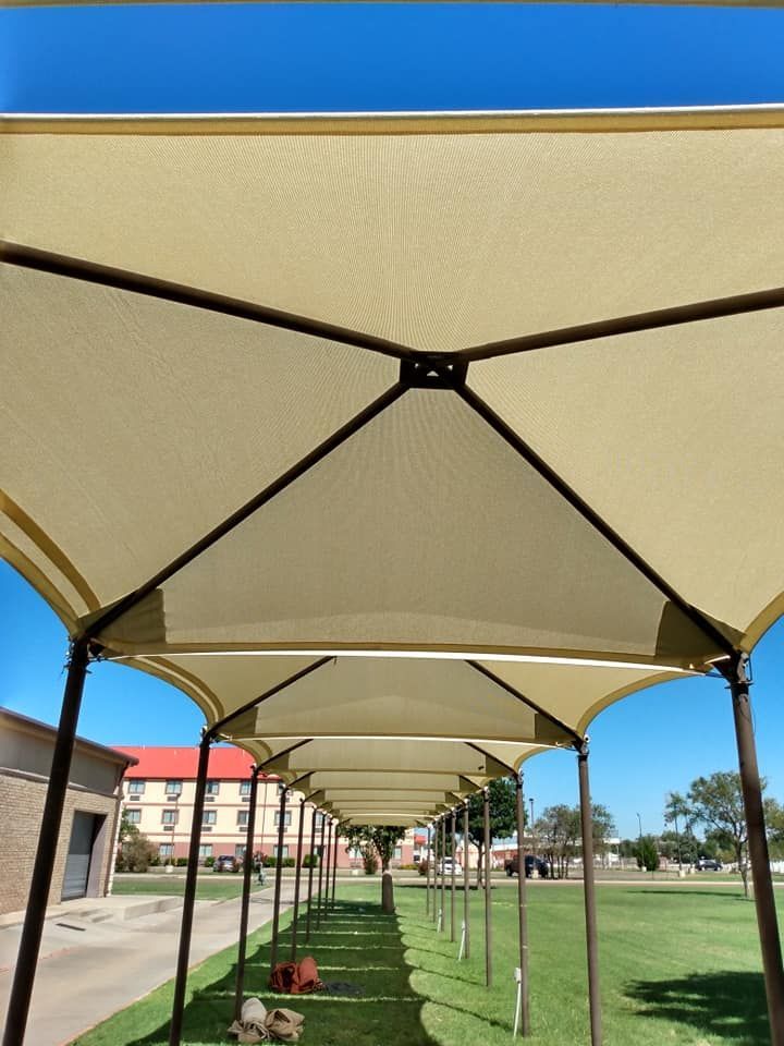A covered walkway with beige shade sails and brown support beams over a green lawn under a blue sky.
