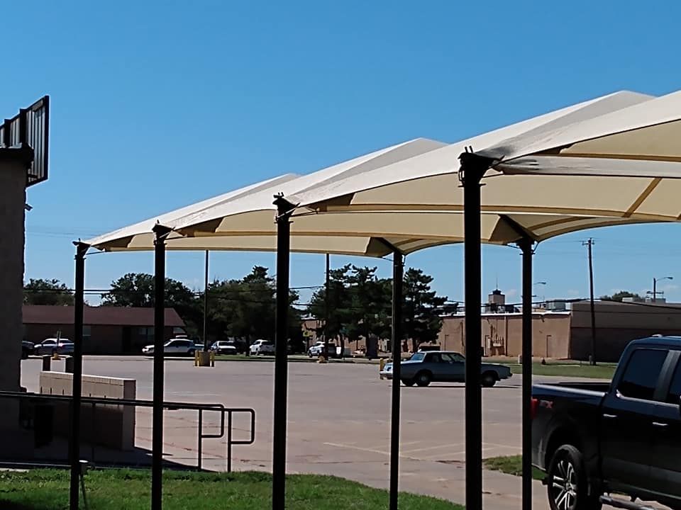 Carport with beige canopies and black poles. Cars parked underneath on a sunny day.