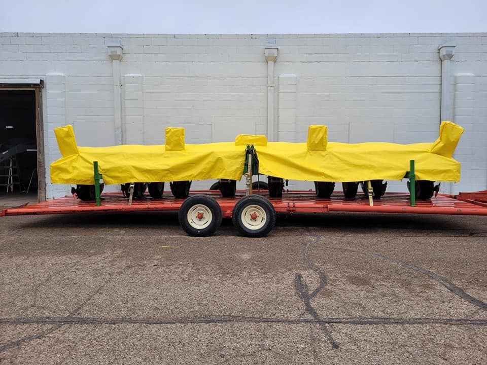 A red trailer with two axles and eight tires carries a covered yellow farm implement in front of a white building.