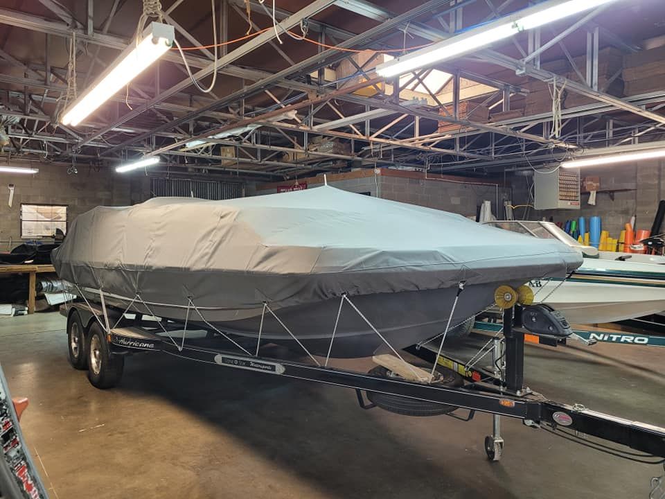 A covered boat on a trailer inside a workshop. Gray boat cover, dark trailer, and ceiling lights visible.