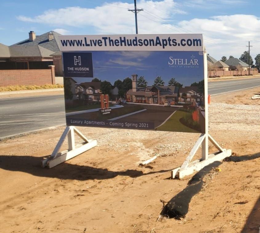 Sign for The Hudson Apartments, website URL and rendering of building, on a stand in a sandy lot.