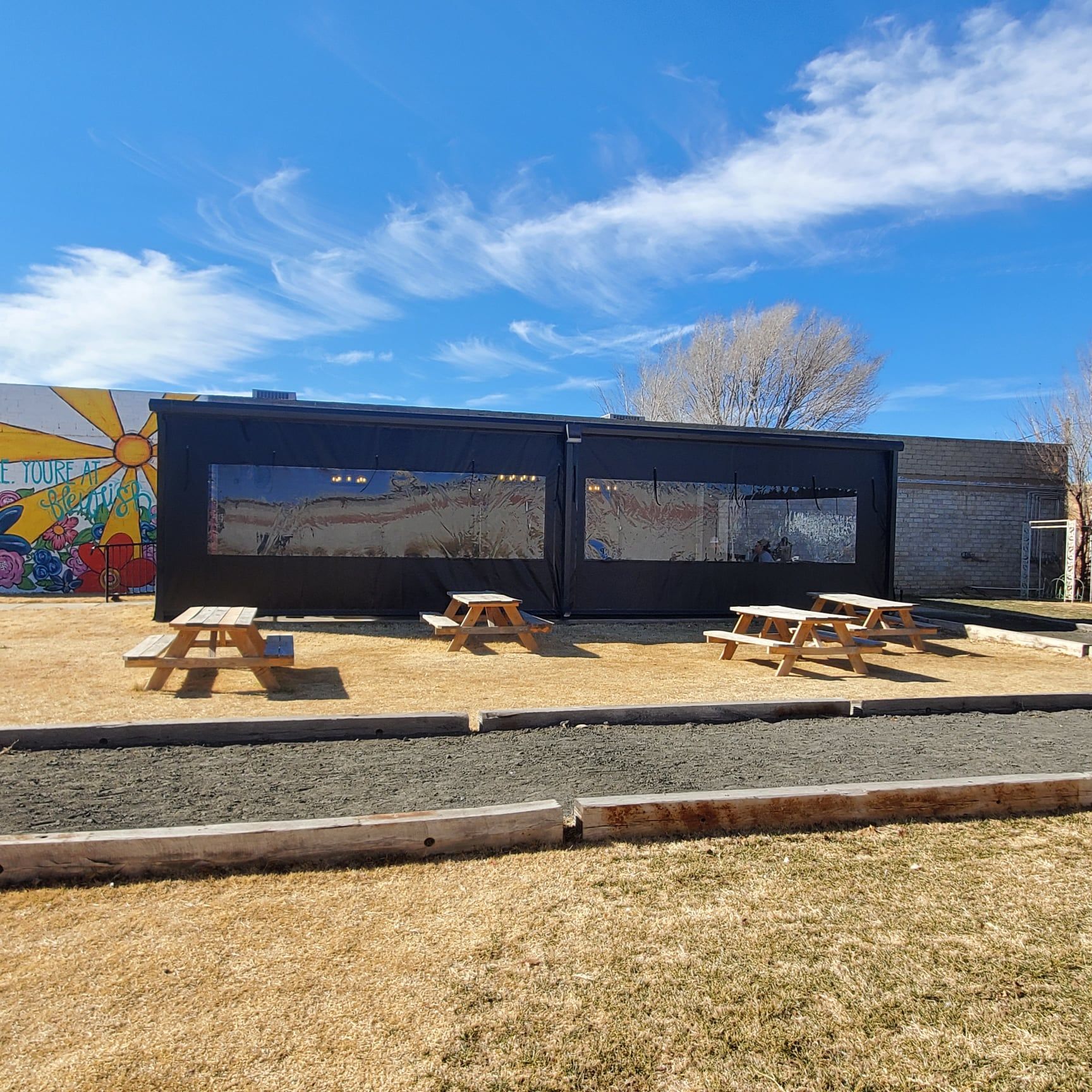 Outdoor dining area with picnic tables in front of a black structure with transparent windows. Blue sky and mural background.