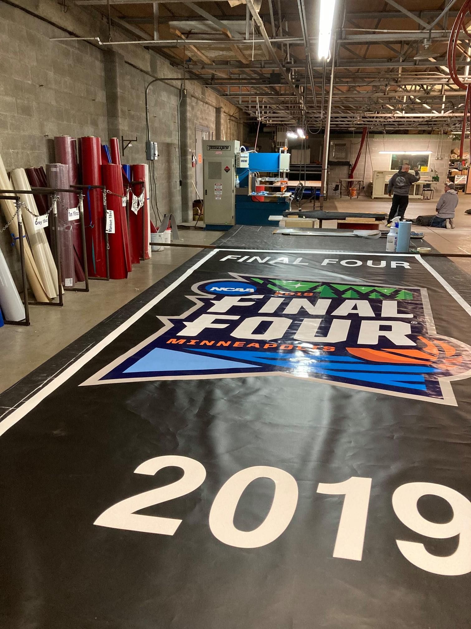 A large 2019 NCAA Final Four floor graphic being worked on in a print shop. Rolls of vinyl are on the left.