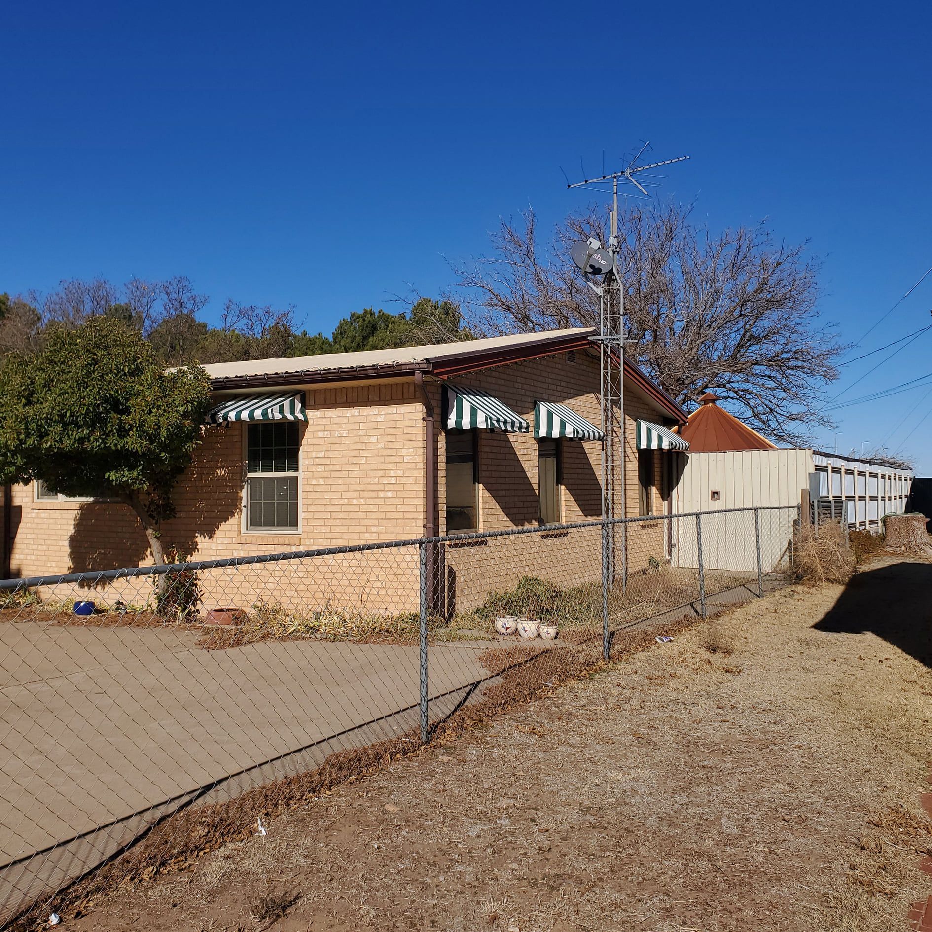 Brick house with green and white striped awnings, chain-link fence, and clear blue sky.