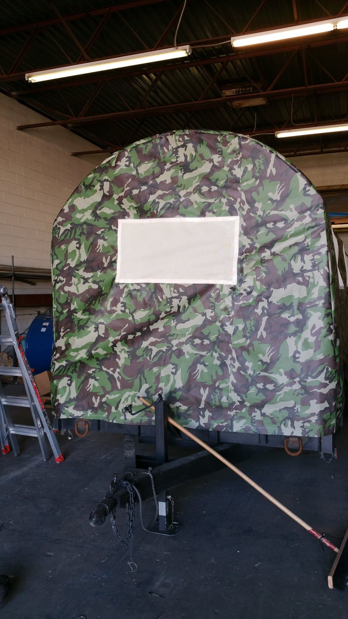 Camouflage-covered, arched structure on a trailer with a rectangular, white window; indoors, industrial setting.
