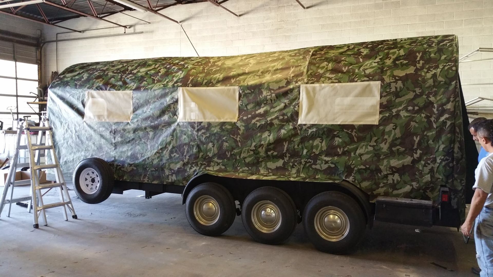 Camouflaged trailer with three axles and four rectangular window openings inside a building.