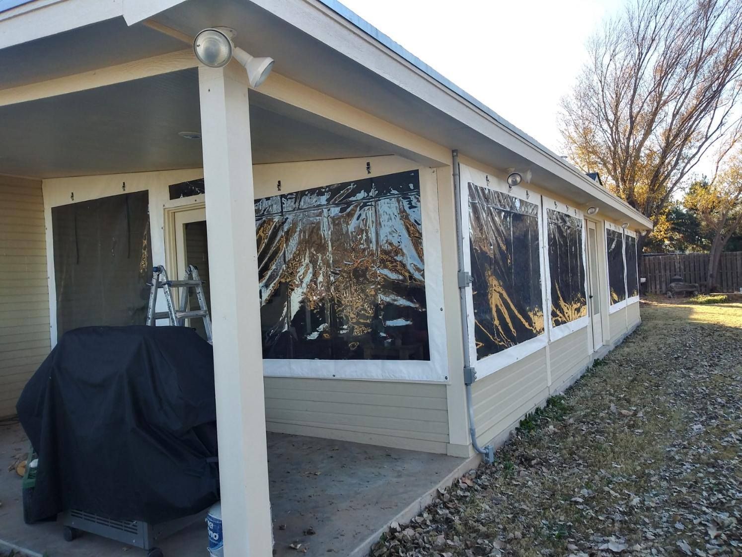 A covered porch with black tarp windows, a grill, and a ladder outside a house.
