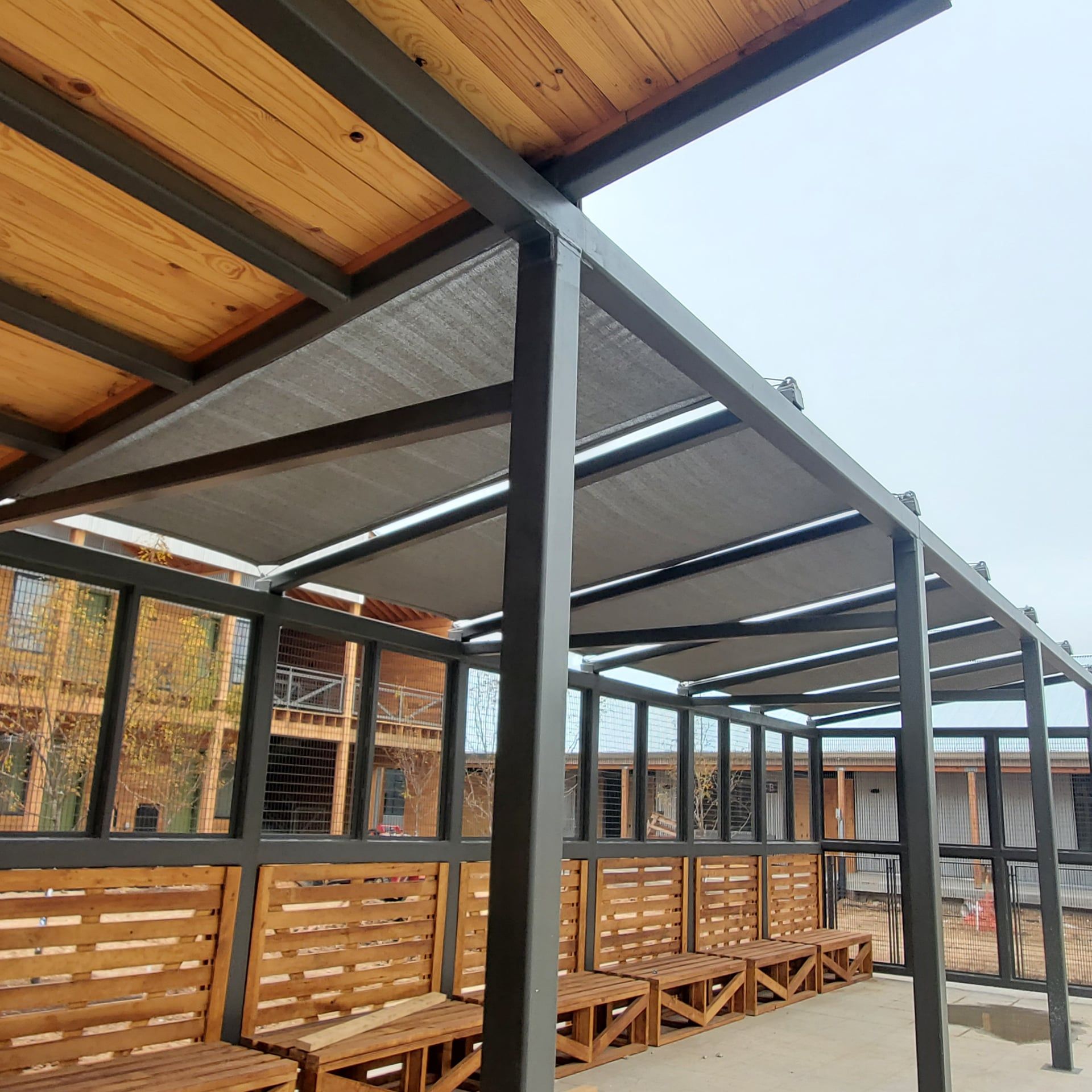 Exterior covered walkway with metal frame, wooden benches, and fabric shade, under a wood ceiling.