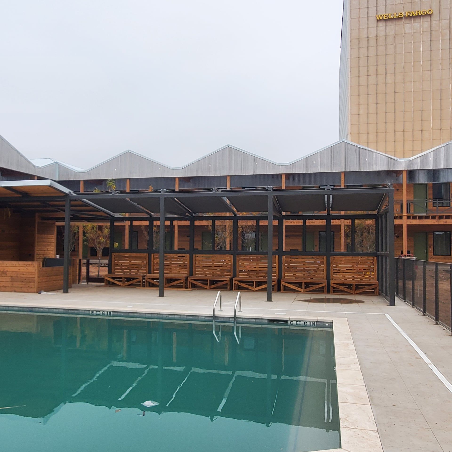 Poolside area with a pool, cabanas, and a tall gold building in the background. Overcast sky.