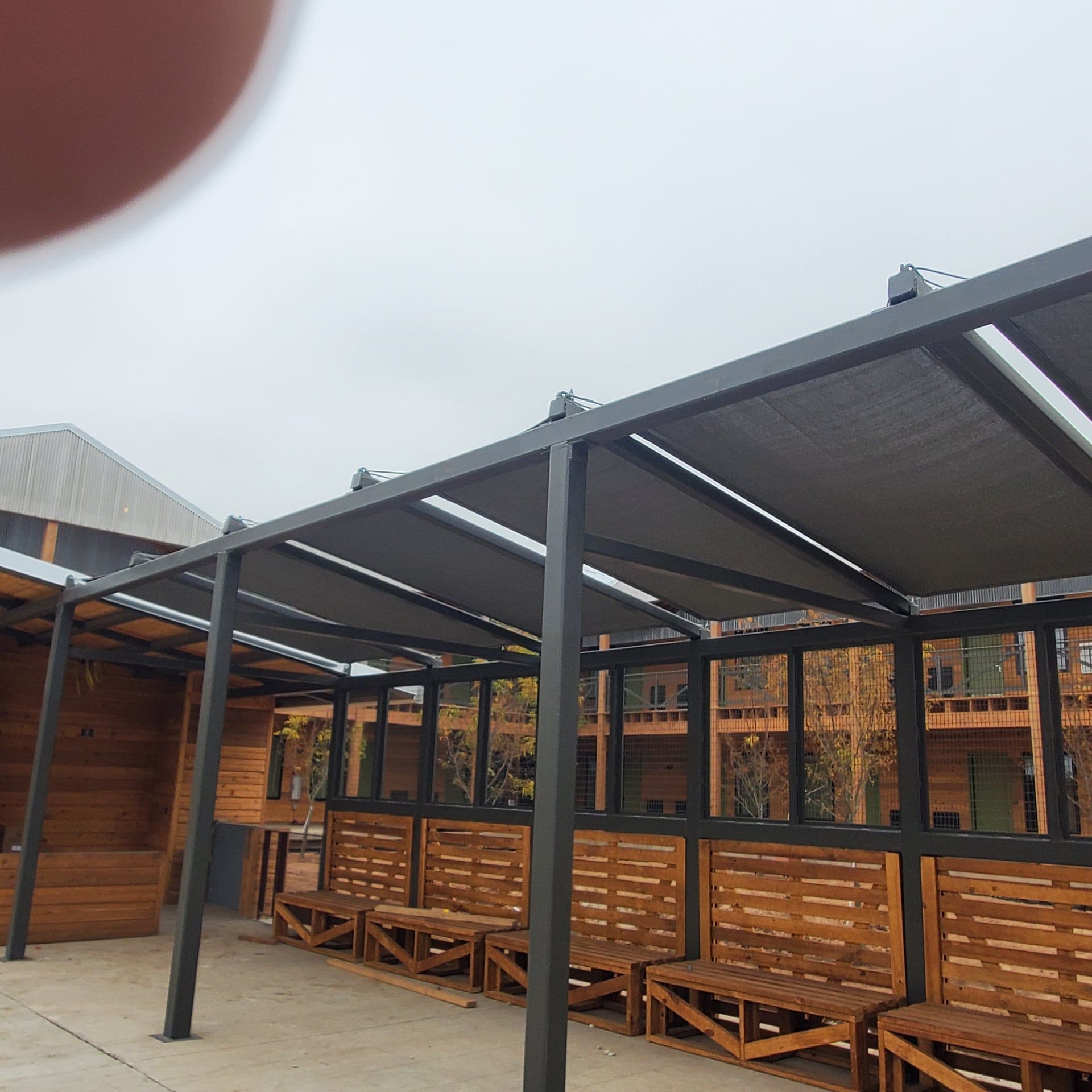 Outdoor seating area with a dark metal pergola, wooden benches, and a cloudy sky.