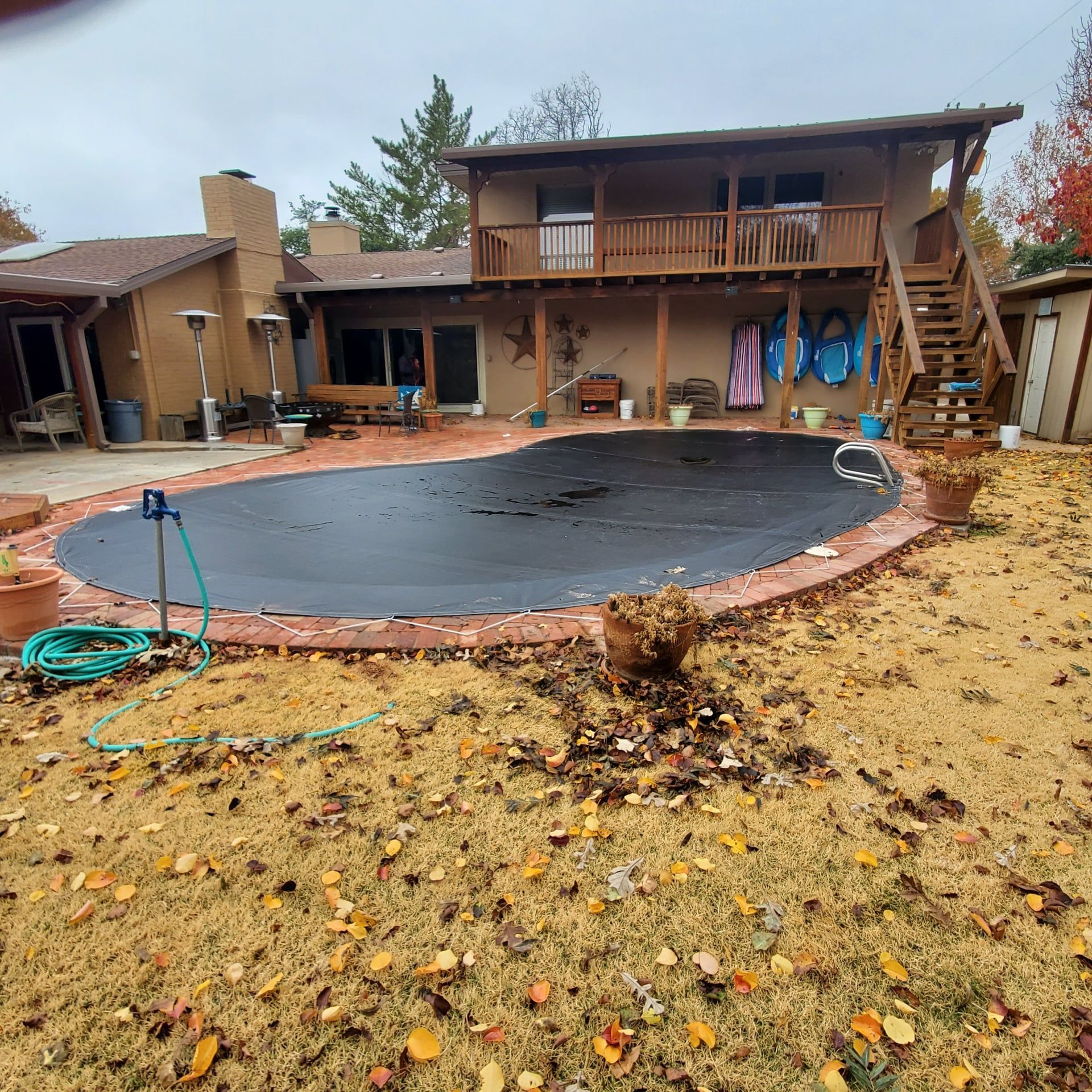 Backyard with a pool covered by a black tarp, surrounded by dry grass and a two-story house.