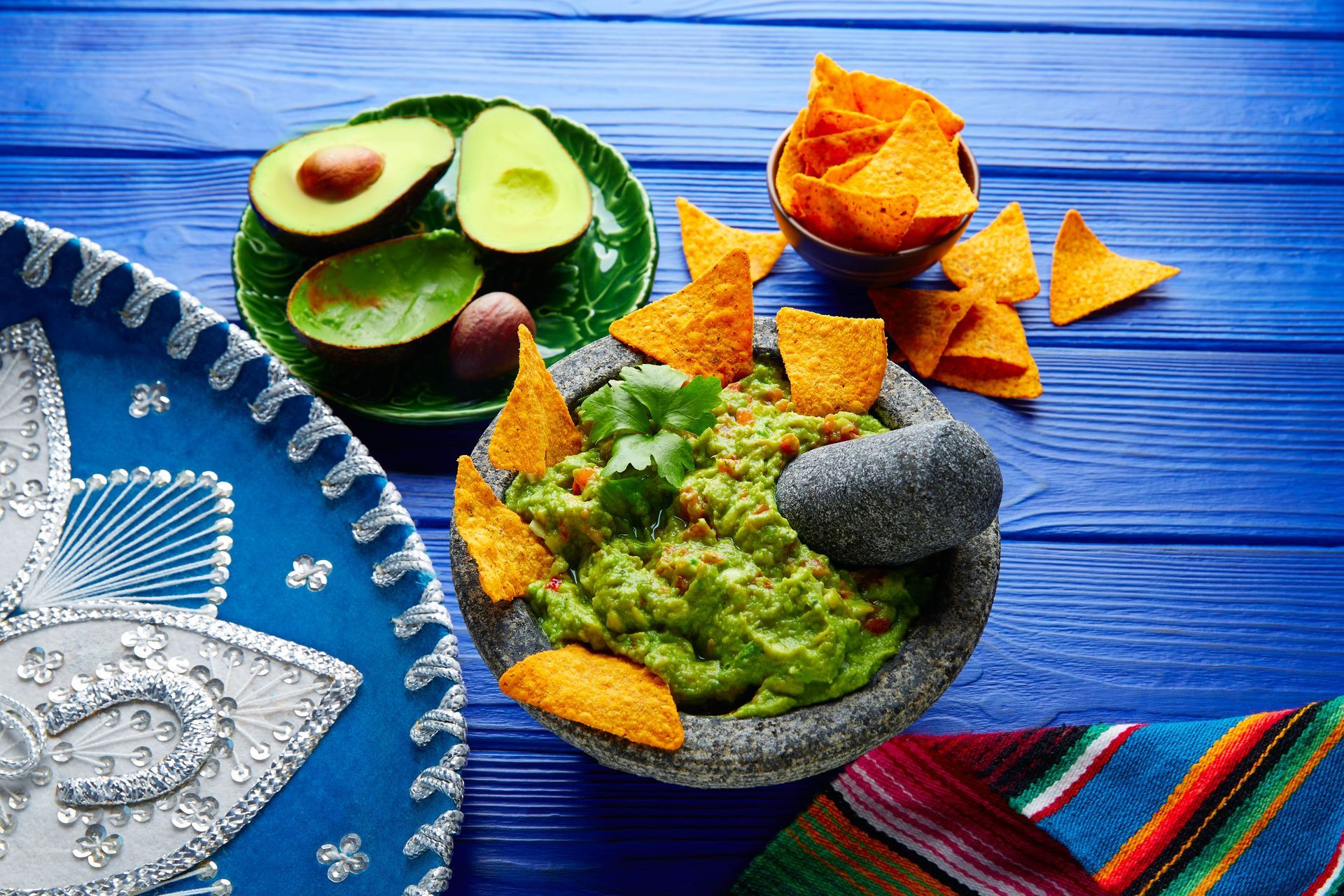 Guacamole in a molcajete with chips, avocados, sombrero, and colorful cloth on a blue wooden surface
