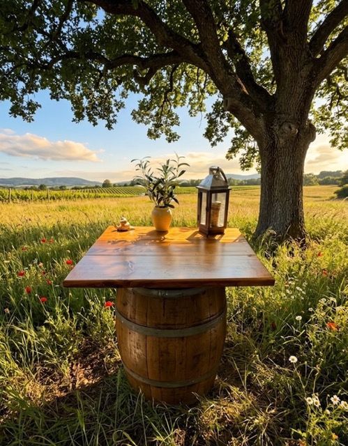 Rustic wooden table on a barrel in a grassy field under a large tree at sunset, with lantern and vase
