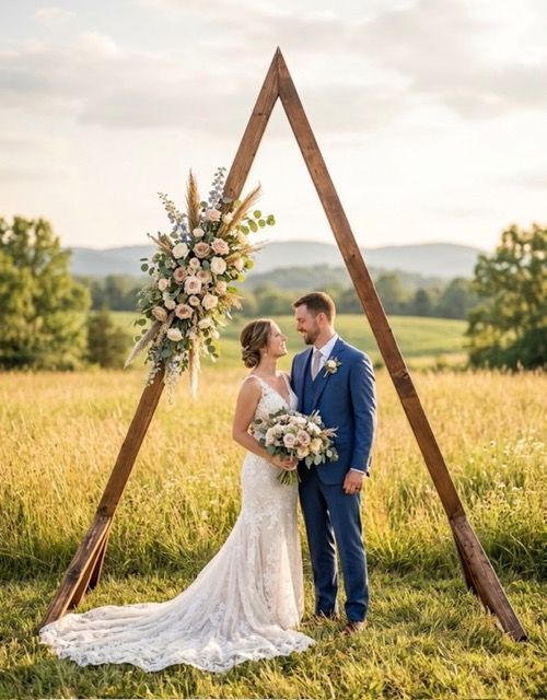 Bride and groom standing under a wooden triangle arch in a wheat field with floral decorations