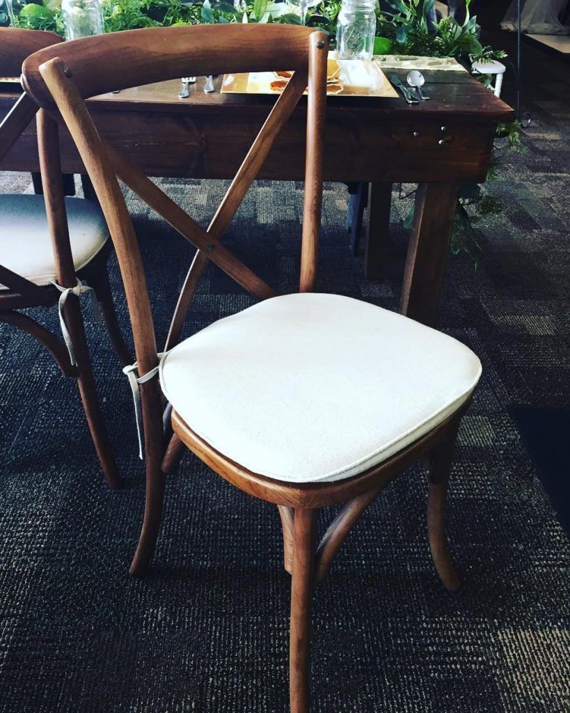 Wooden chair with a white cushion and an X-shaped back, set in front of a table.