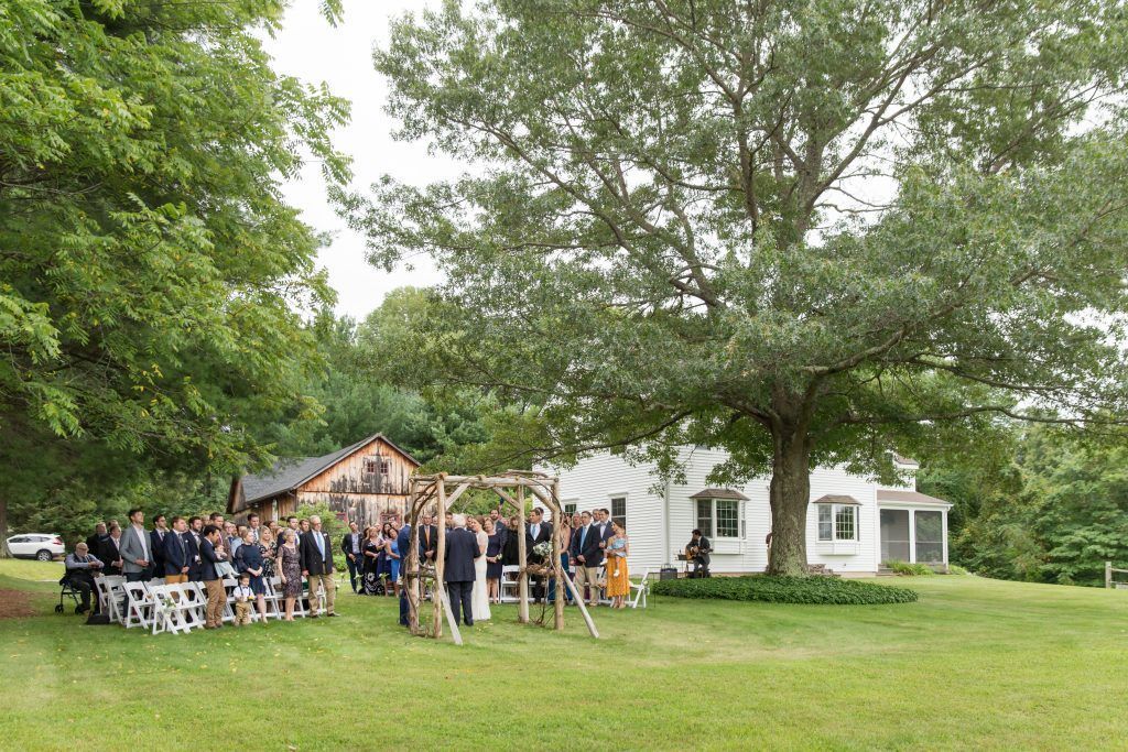 Wedding ceremony outdoors, guests seated on benches, couple under wooden arch, white house and tree in background.
