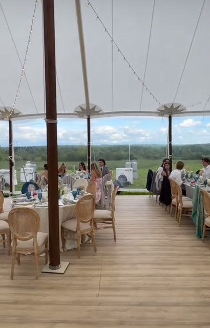 Wedding reception inside a tent, overlooking a field. Tables set with guests. Wooden floors, tan chairs, white tent.