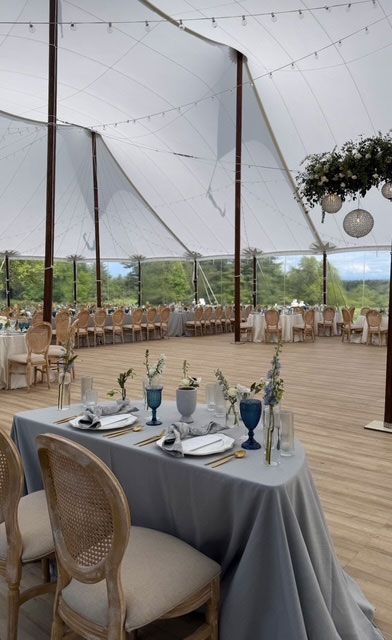 Wedding reception tent with tables set for guests, wooden floors, string lights, and floral decorations.