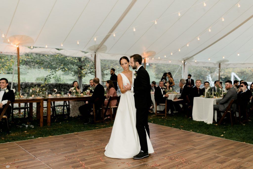 Bride and groom dance on a wooden dance floor in a tent, guests at tables.