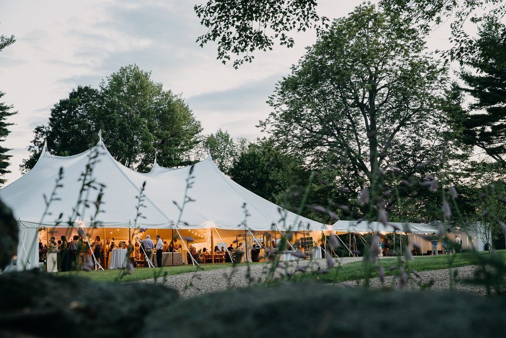 White tent lit up at dusk, hosting a social gathering. Trees and foliage surround the event.