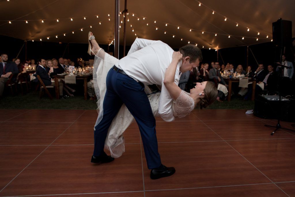 Couple dancing at wedding reception. Groom dips bride on a wooden dance floor under tent with string lights.