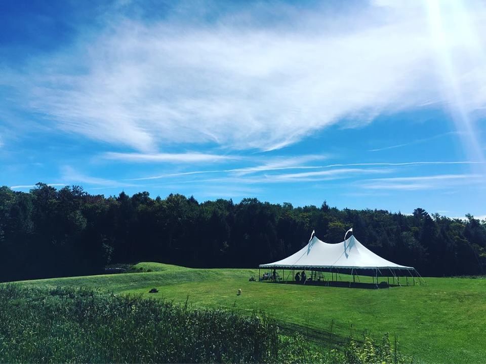 A white tent on a grassy field, with a forest backdrop under a blue sky.