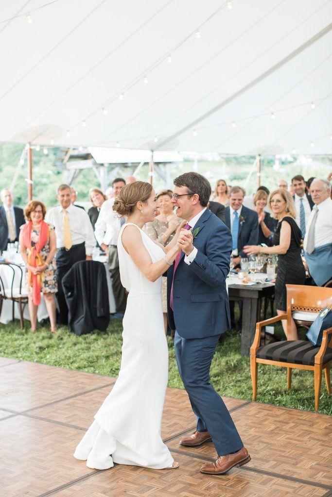 Bride and groom dance at a wedding reception under a tent, smiling. Guests watch.