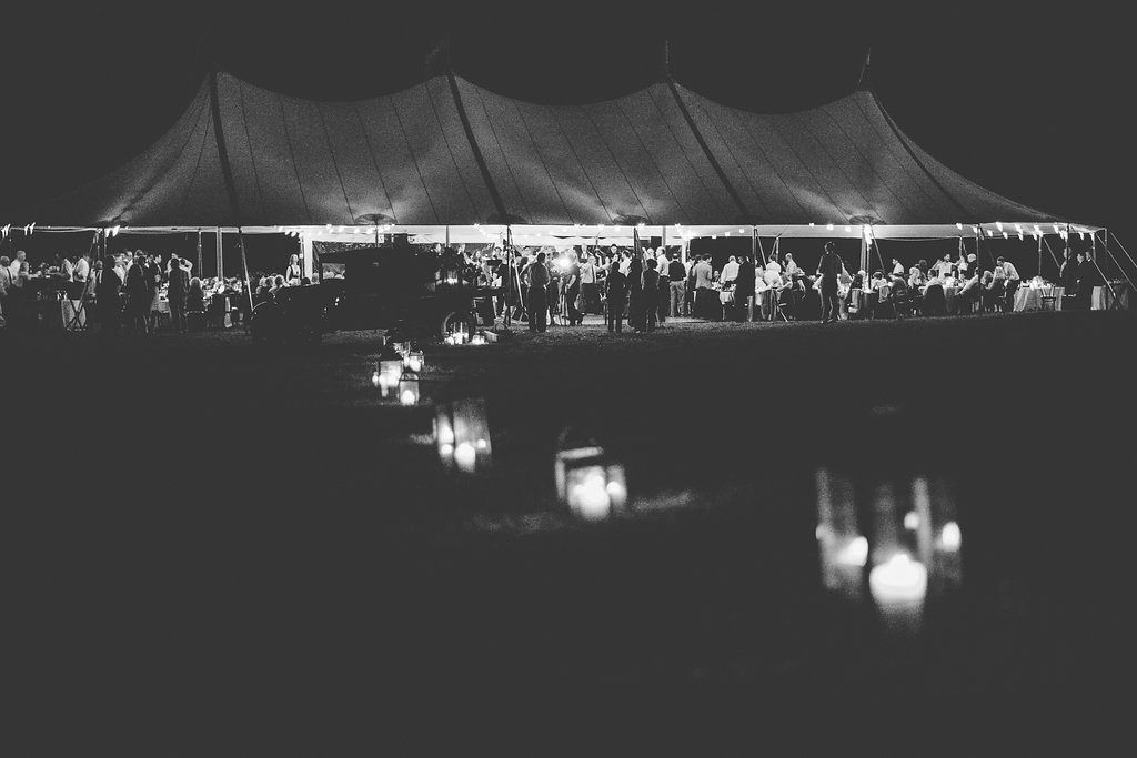 Large outdoor tent at night with lights on, hosting a gathering; water in front reflects the lights.