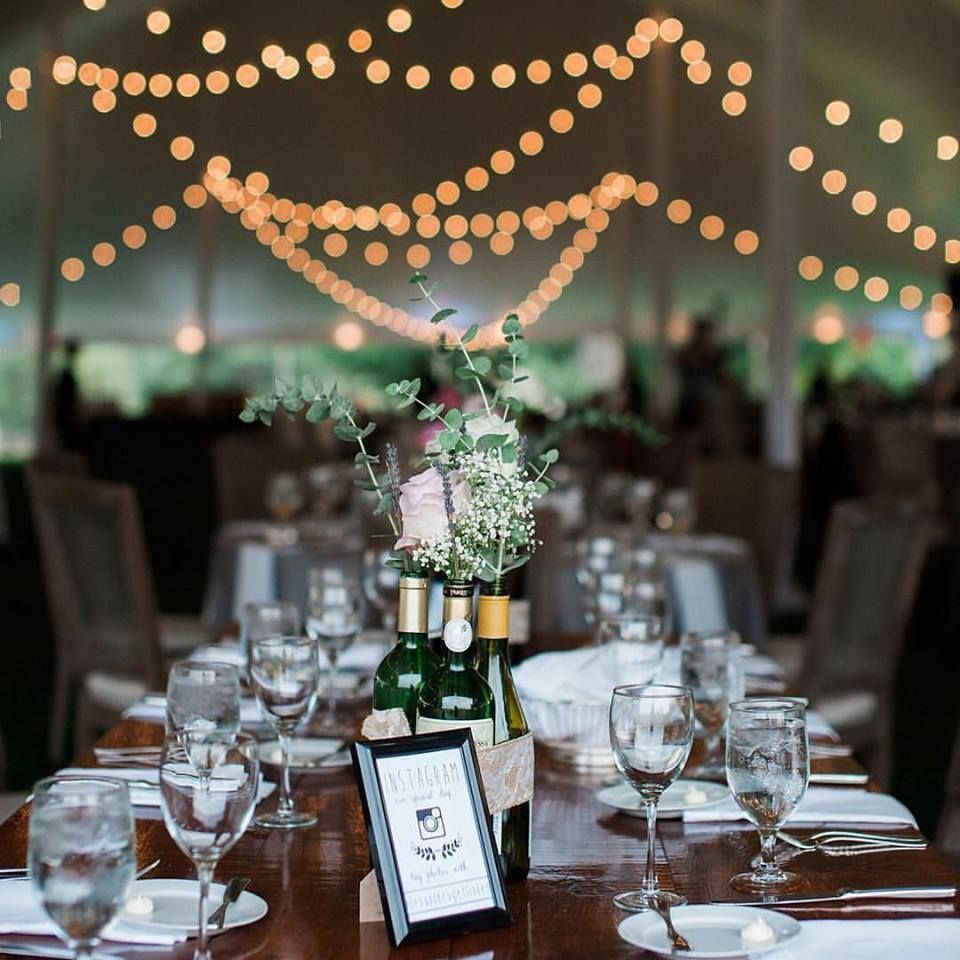 Wedding reception table setting with floral arrangement and string lights in a tent.