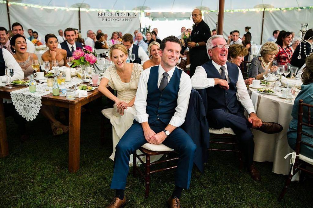 Wedding reception: Guests seated at tables under a tent; man in blue suit and vest smiles.